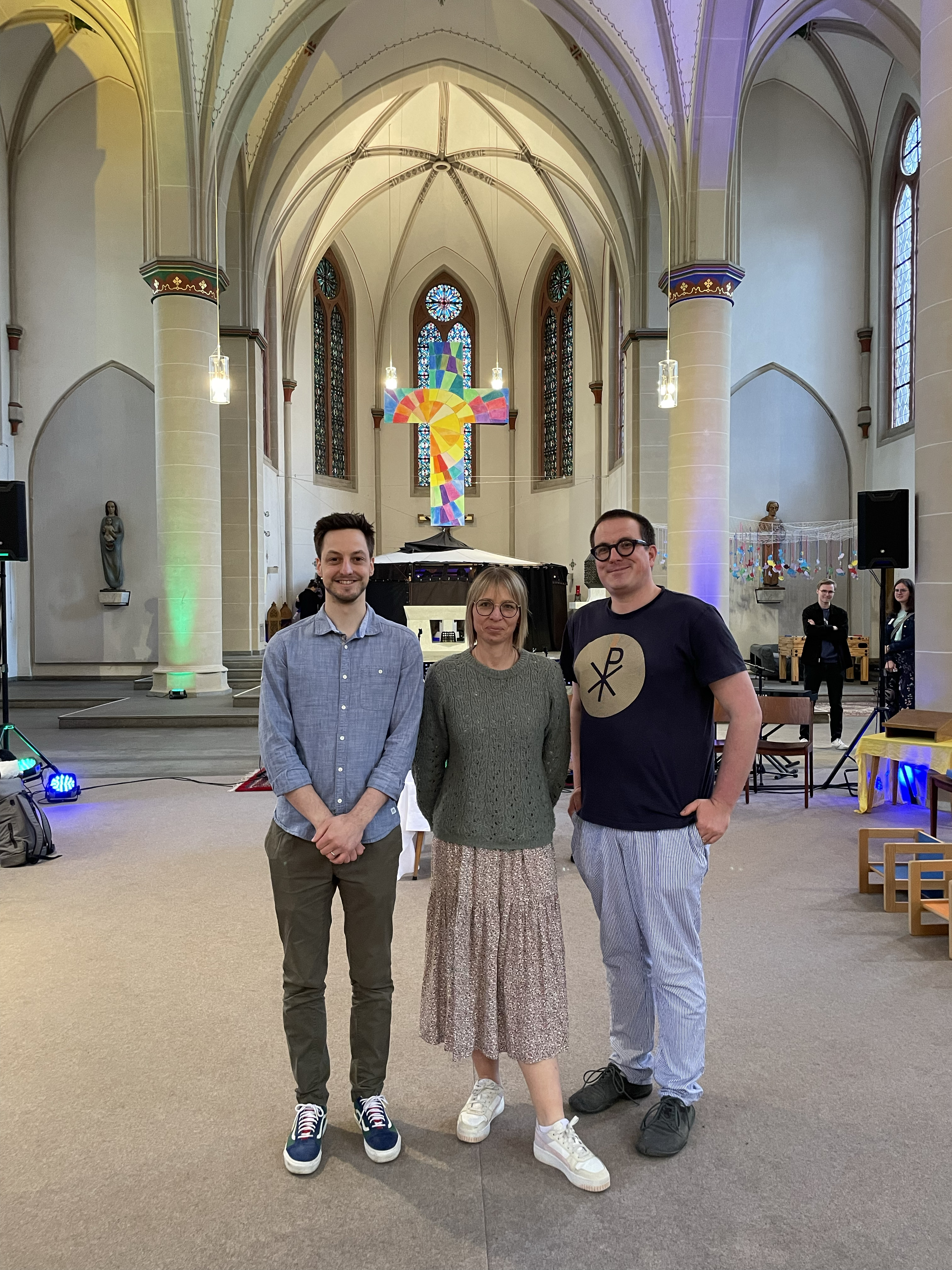 Drei Personen stehen in einer Kirche im Hintergrund der Altar, links ein Mann, rechts ein Mann, in der Mitte eine Frau