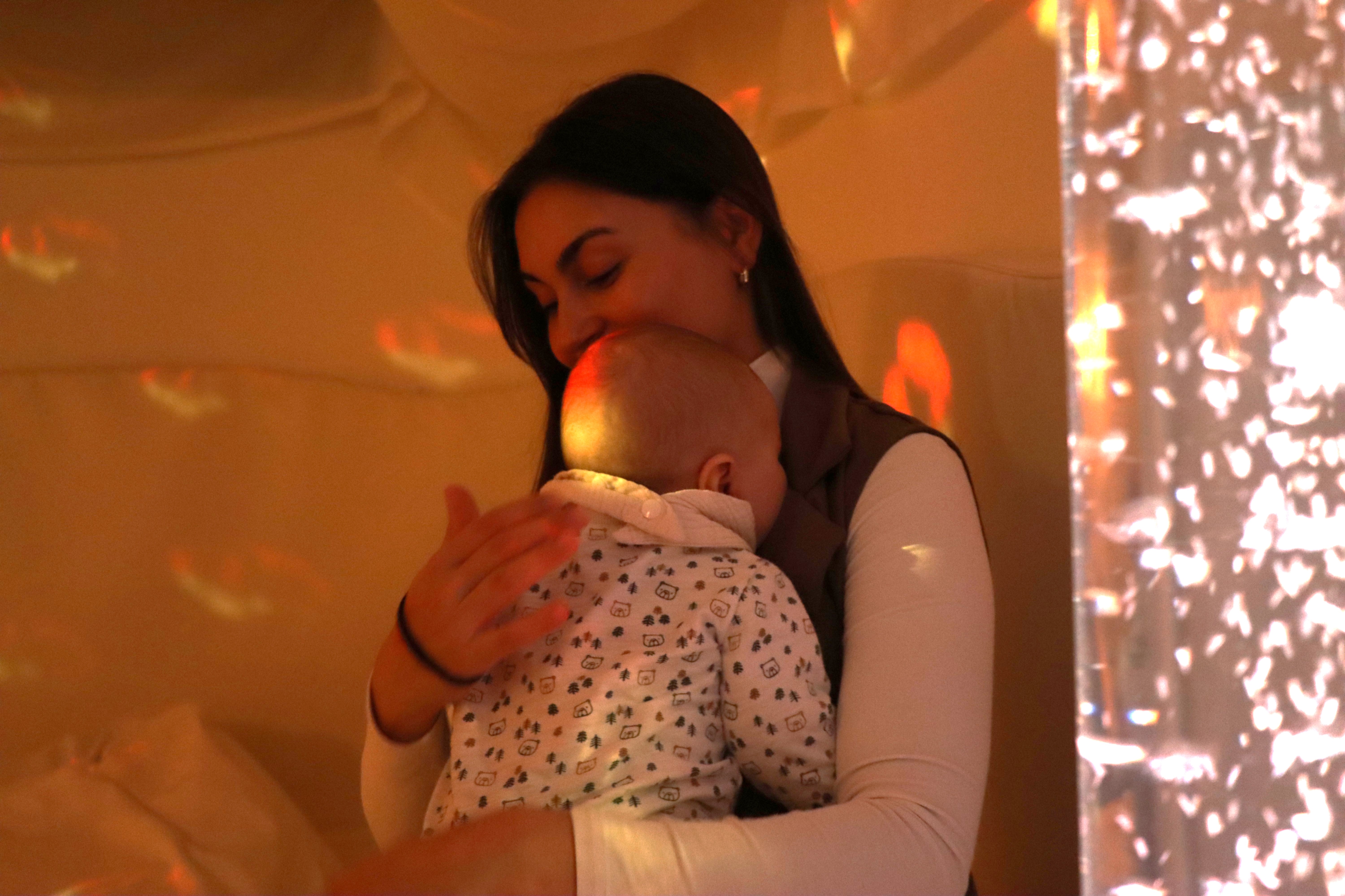 Eine junge Frau sitzt in in einem Raum mit warmem, orangenen Licht. Sie hält ihr Baby auf dem Arm, streicht ihm über den Rücken. Am rechten Bildrand ist eine sprudelnde Wassersäule aus Plexiglas zu sehen, die das Licht in bunten Mustern durch den Raum und über das Gesicht der Frau wirft. 