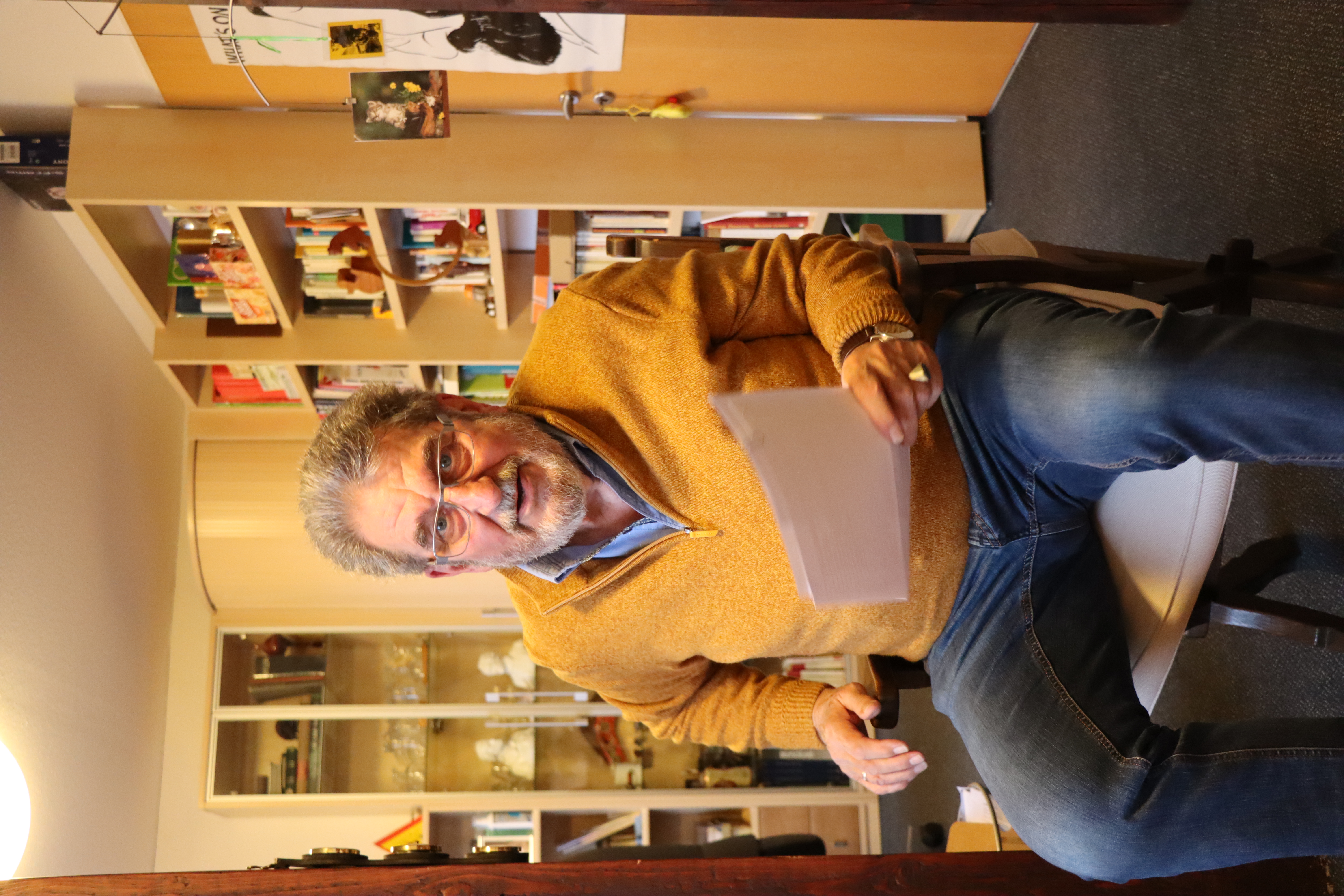 Ein Mannsitzt auf einemStuhl, hinter ihm im Raum Regale gefüllt mit Gegenständen