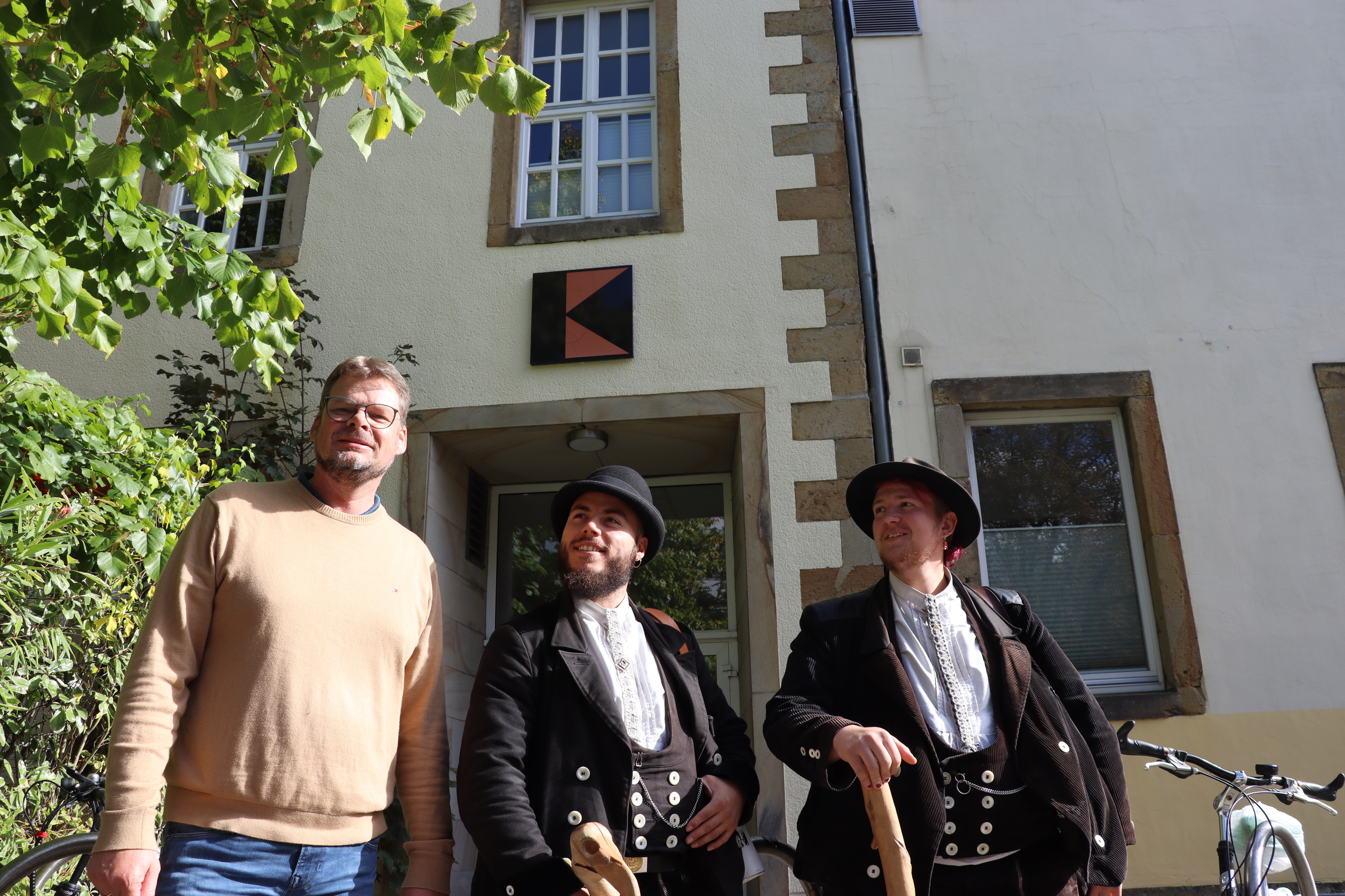 Stephan Düing steht mit beiden Gesellen vor dem Osnabrücker Kolpinghaus. Die jungen Männer tragen traditionelle Kleidung in schwarz, halten die Wanderstöcke in den Händen. 