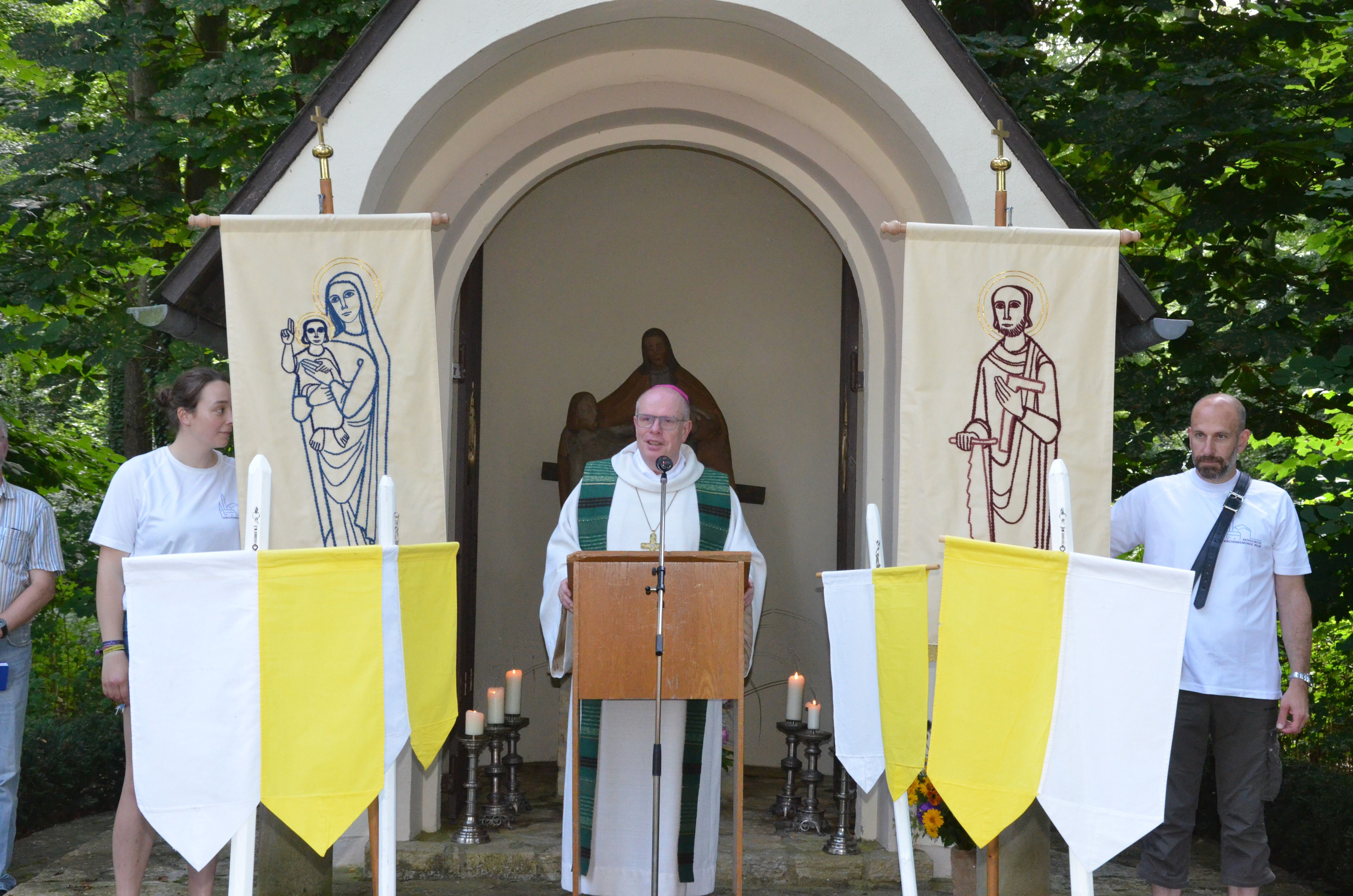 Weihbischof Wübbe predigt vor einer Kapelle, neben ihm stehend zwei Bannerträger