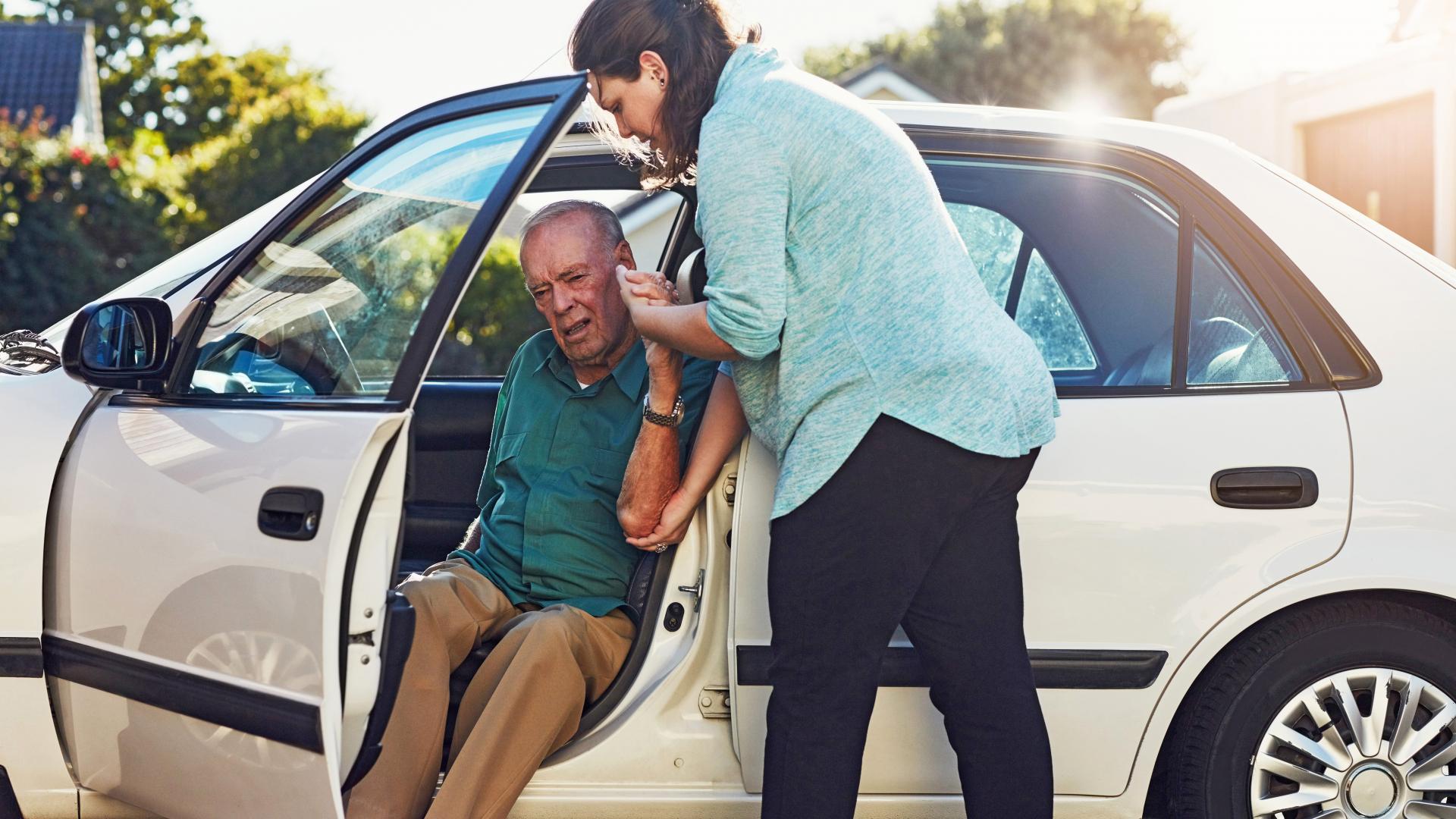 Eine junge Frau hilft einem älterem Mann, vom Fahrersitz aus aus dem Auto auszusteigen. 