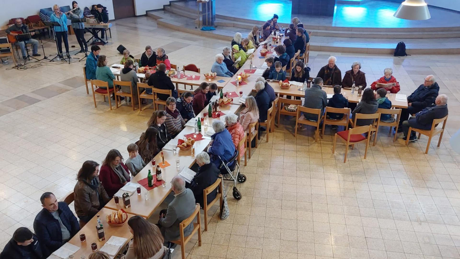 Gemeindemitglieder sitzen zur Agape-Feier an Tischen, die zu einem Kreuz aufgestellt sind