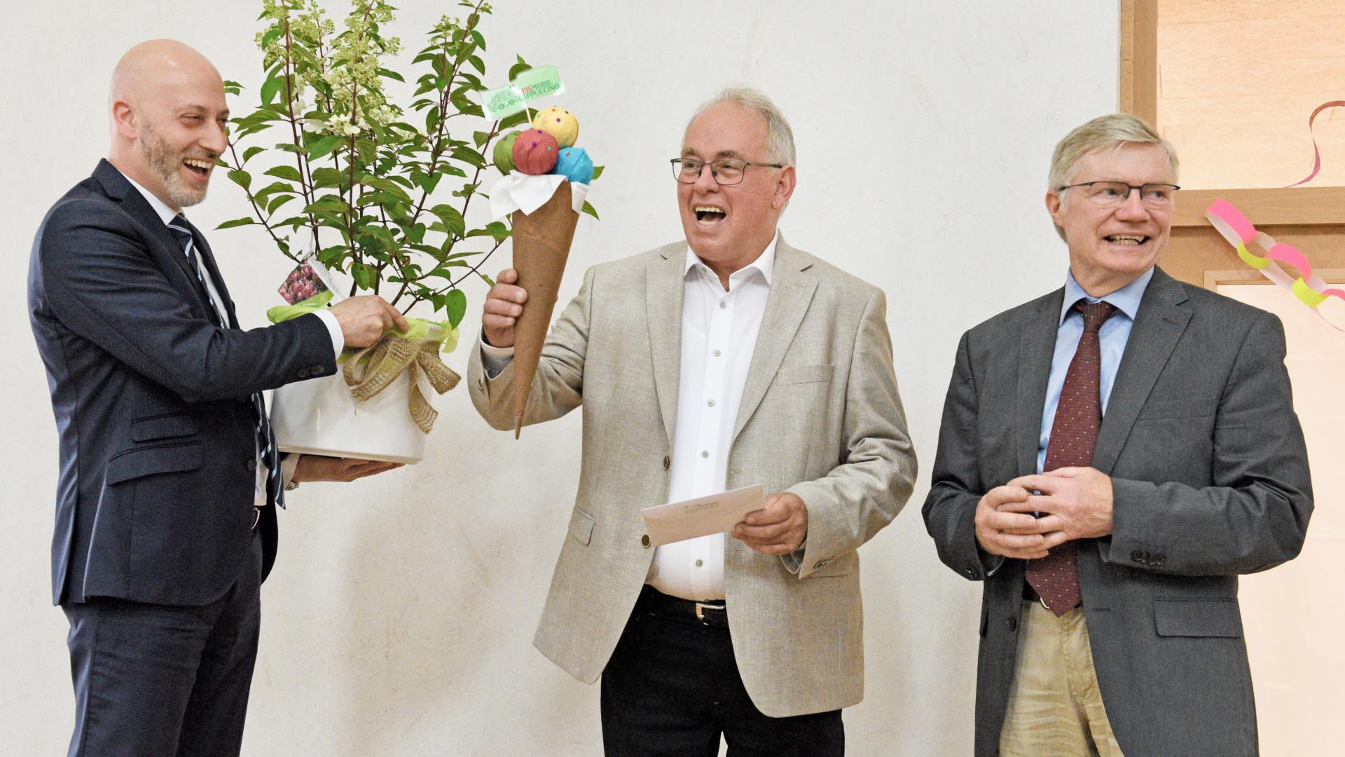 Drei Männer, einer übergibt eine Pflanze, ein anderer hält ein großes Eis aus Papier in den Händen.