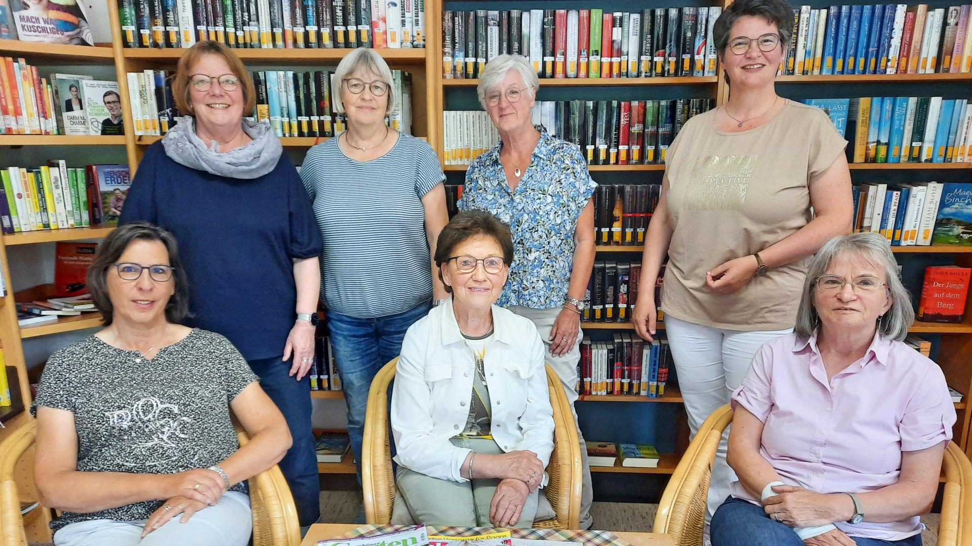 Sieben Frauen stehen und sitzen in zwei Reihen vor einem gefüllten Bücherregal