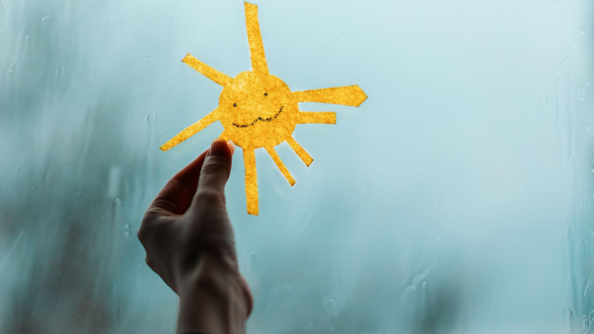 Eine Hand hält ein leuchtendes Sonnenpapier vor ein verregnetes Fenster