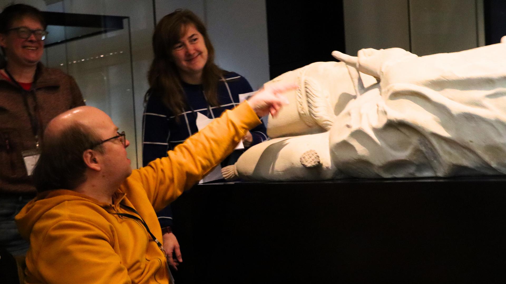 Drei Menschen stehen im dunklen Raum des Osnabrücker Diözesanmuseums. Ein Mann sitzt im Rollstuhl, vor ihm liegt eine große steinerne Statue. Sie reicht dem Mann jedoch über den Kopf. Da er im Rollstuhl sitzt kann er die Statue nicht sehen. Die beiden Frauen, die neben ihm stehen, können die Statue problemlos sehen - schauen auf sie herab. 