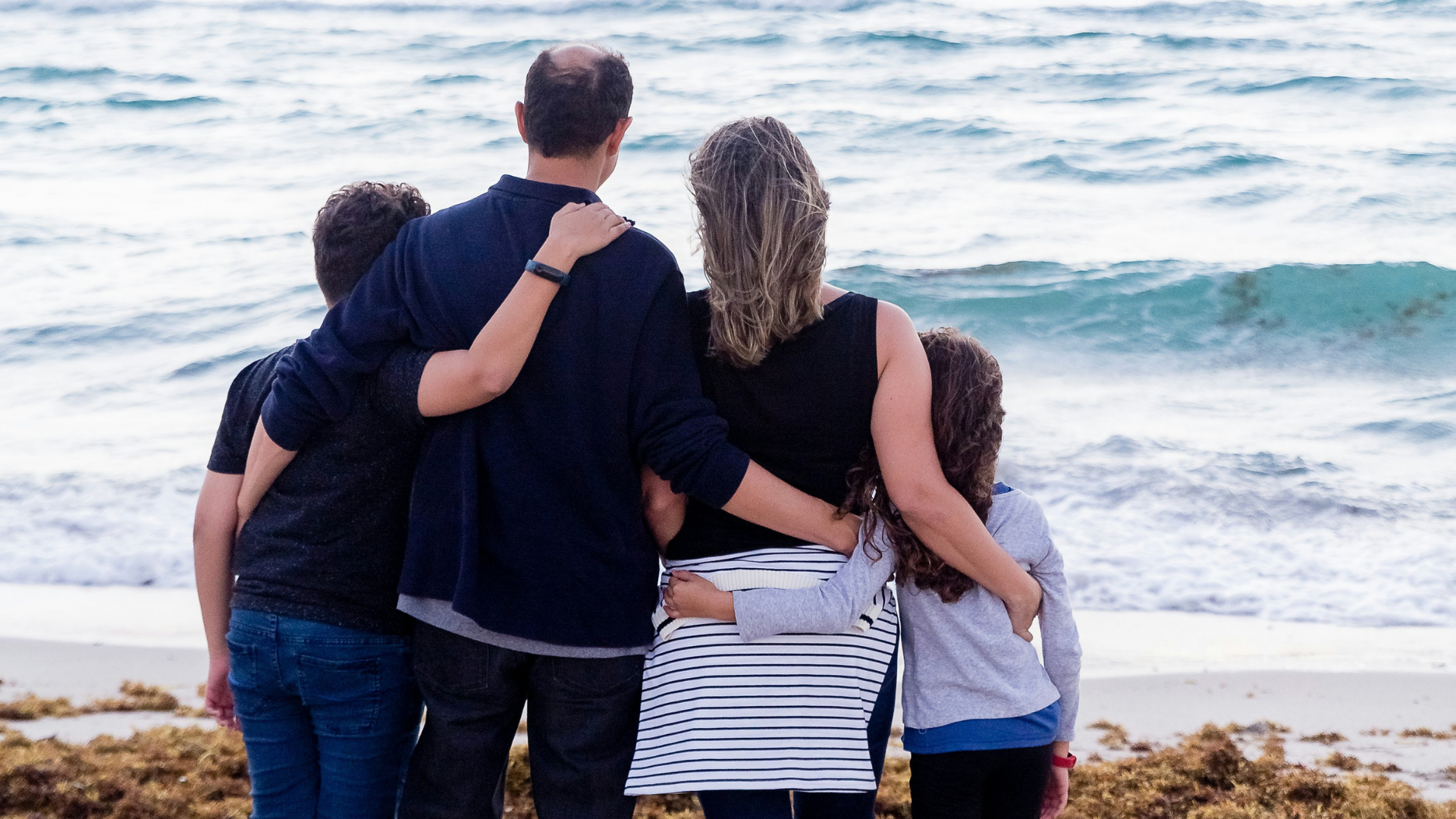 Rückansicht einer sich umarmenden Personen (Sohn, Vater, Muttern Tochter) im Hintergrund das Meer