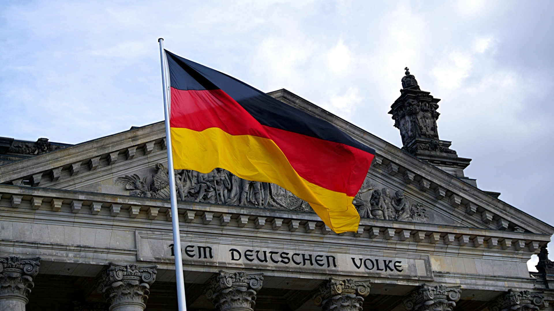 Vor dem Reichstagsgebäude in Berlin weht eine große deutsche Flagge im Wind. Auf dem Gebäude ist die Inschrift ‚Dem Deutschen Volke‘ zu sehen. Der Himmel ist leicht bewölkt.
