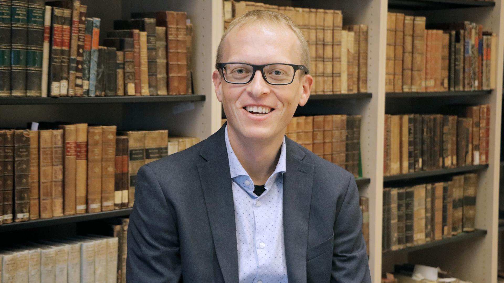 Ein Mann lächelt vor einem Bücherregal sitzend in die Kamera.