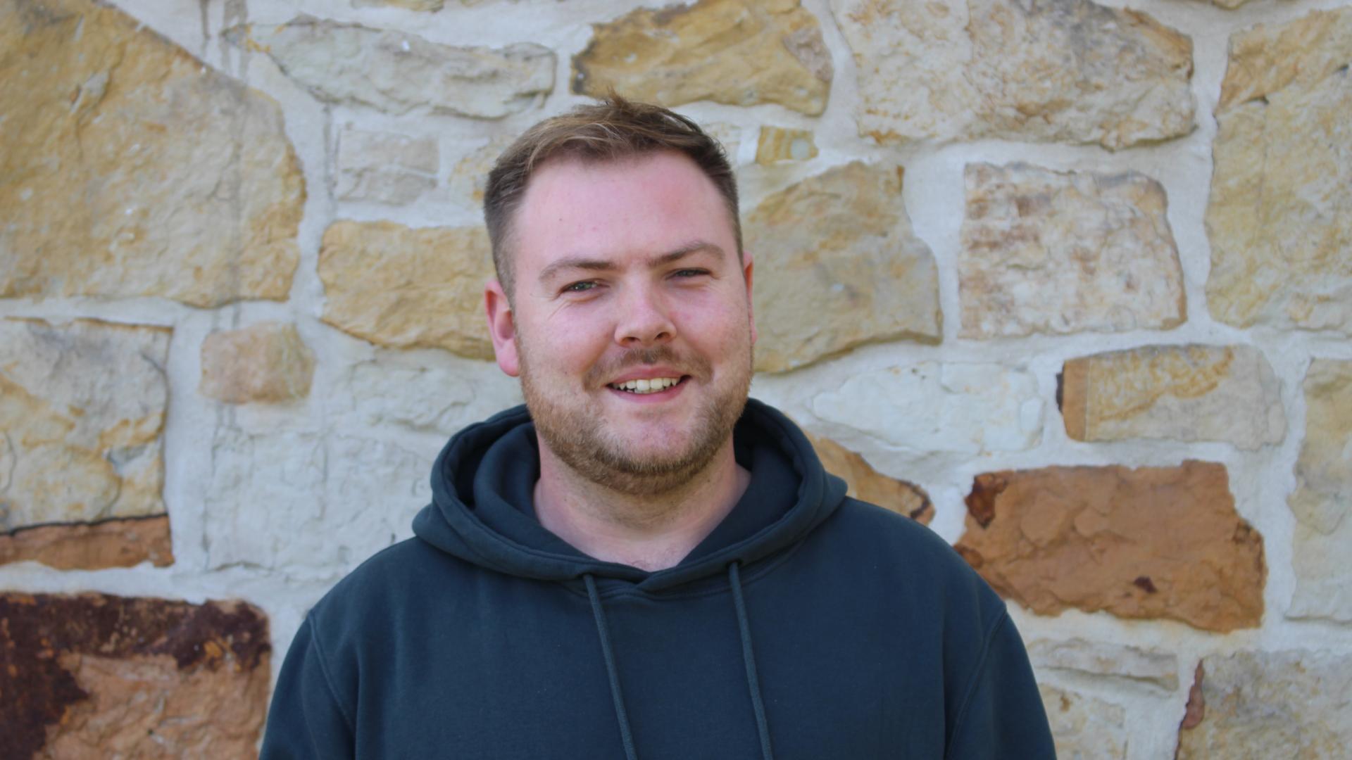 Ein Mann mit schwarzem Pullover vor einer Sandsteinwand