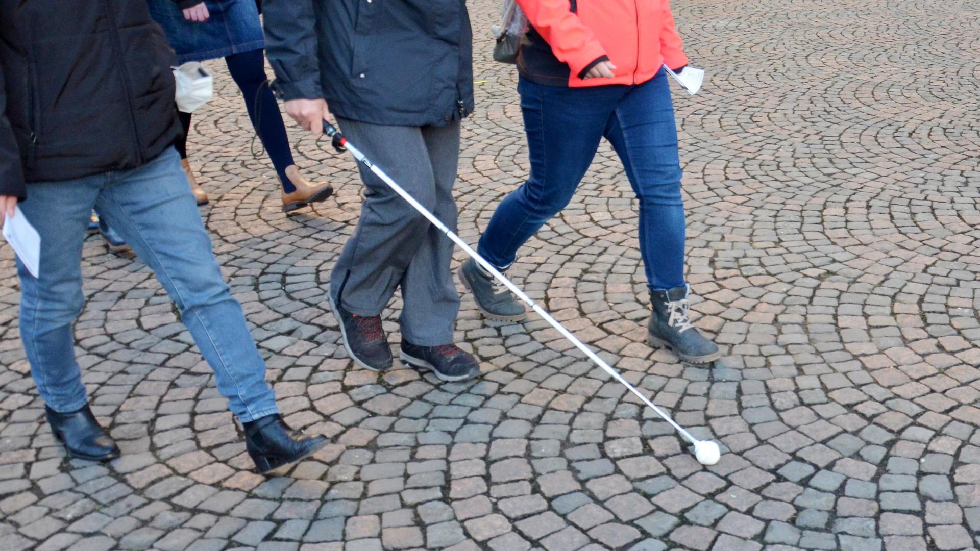 Zu sehen sind die Beine von drei Menschen, eine der Personen geht mit einem Blindenstock.