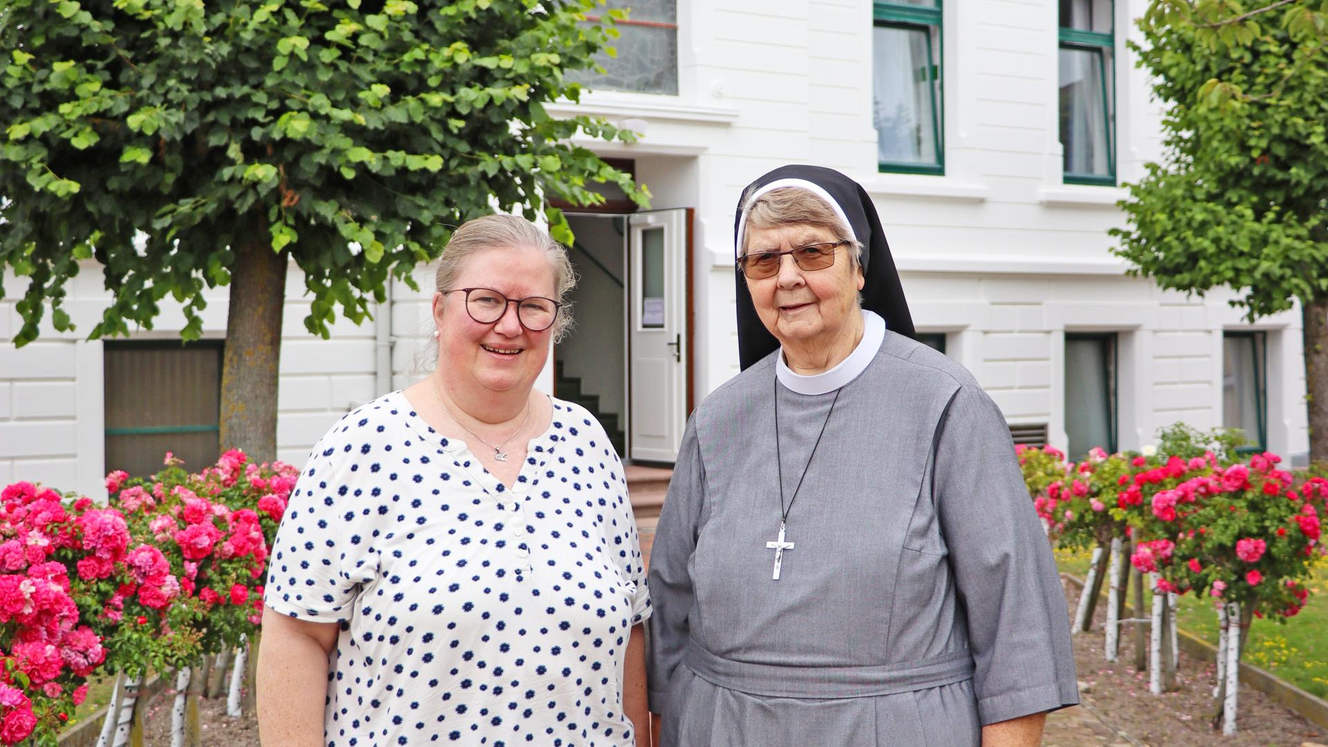 Eine Frau steht neben einer Nonne vor einem weißen Haus