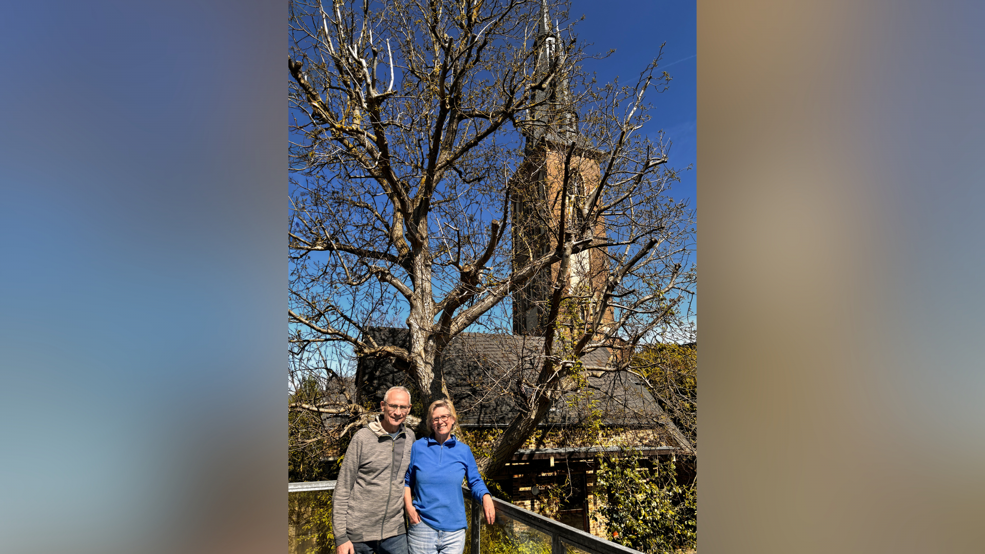Zwei Menschen stehen auf Balkon, hinter ihnen ragt Kirchturm in blauen Himmel. 