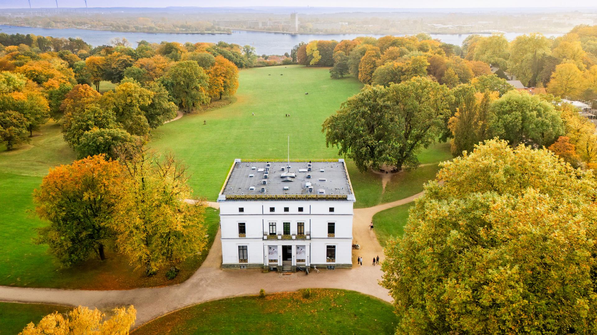 Zu sehen ist ein weißes Gebäude inmitten einer Parkanlage mit Rasen und Bäumen.