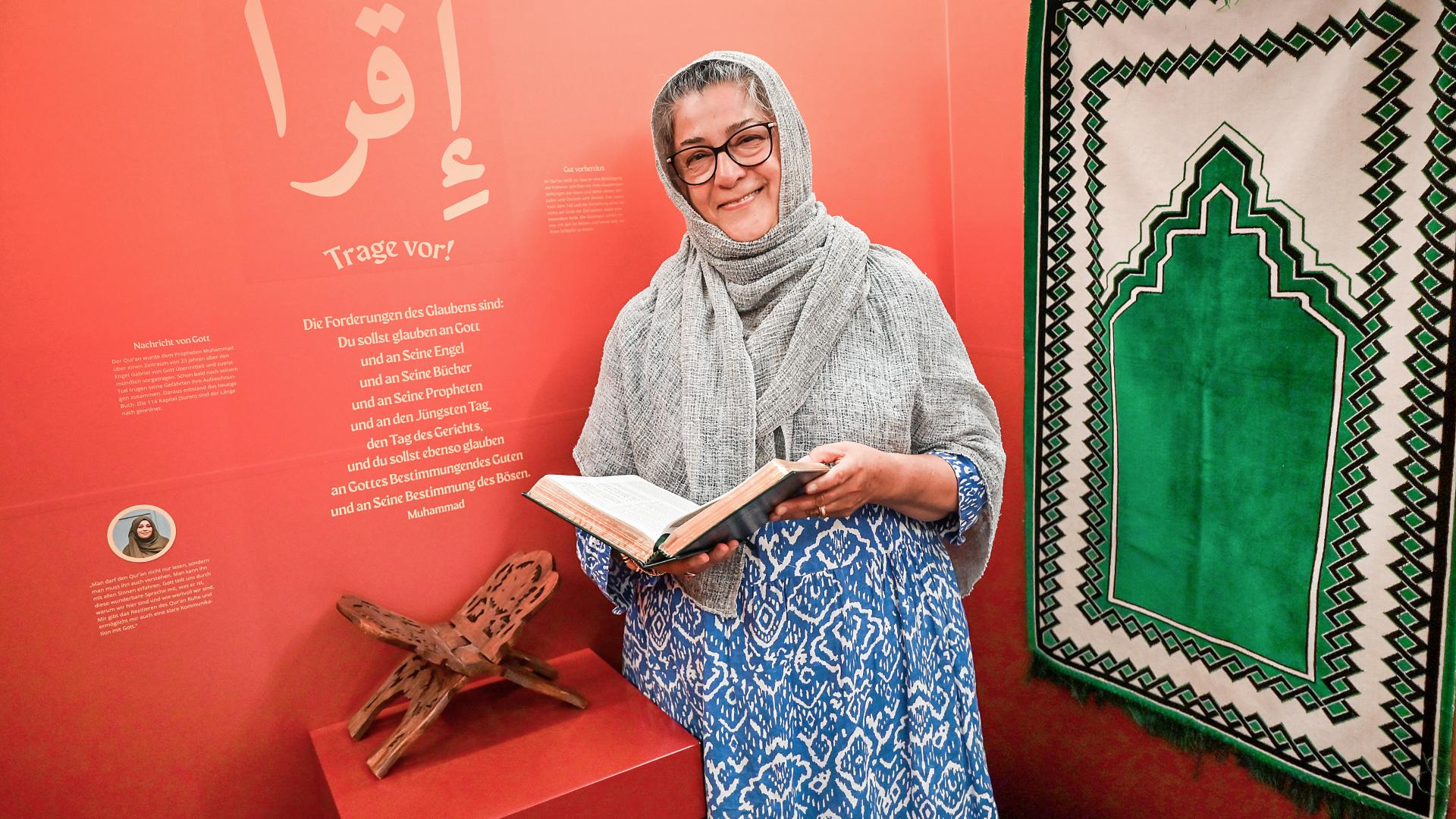 Hamideh Mohagheghi steht vor einer roten Wand mit Zitaten aus dem Koran, ein grüber Teppich hängt an der Wand. Sie lächelt, trägt ein Kopftuch und blättert im Koran. 