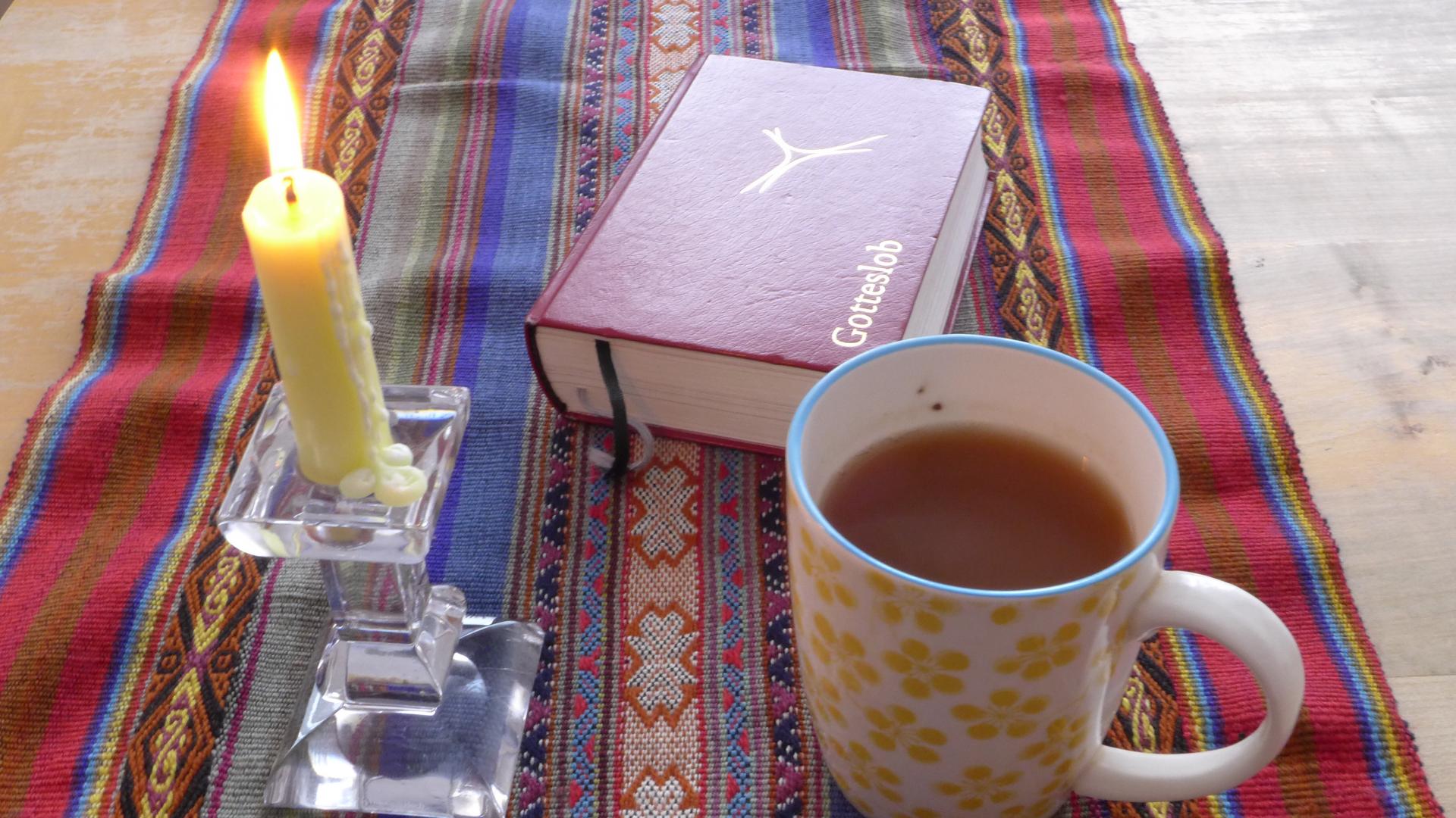 Zu sehen ist ein Gotteslob, eine Kerze und eine Tasse mit Tee auf einem Tisch. 